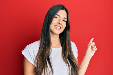 Young brunette woman wearing casual white tshirt over red background with a big smile on face, pointing with hand and finger to the side looking at the camera. 