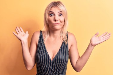 Sarışın, çizgili tişört giyen genç ve güzel bir kadın izole edilmiş sarı arka planda duruyor. Hiçbir şeyden habersiz, kolları açık, hiçbir fikri yok ve şüpheli bir yüzü var..