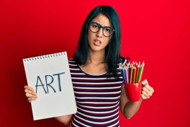 Güzel Asyalı genç bir kadın elinde sanat defteri ve renkli kalemler tutuyor. Hiçbir şeyden habersiz ve kafası karışmış ifadeler. şüphe kavramı. 