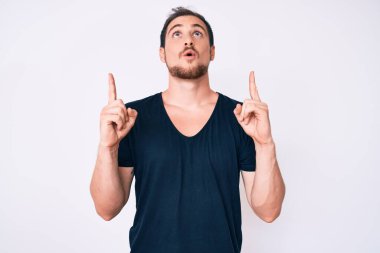 Gündelik giysiler giyen genç yakışıklı adam şaşırmış ve şaşırmış bir şekilde yukarı bakıyor ve parmaklarıyla işaret ediyor ve kollarını kaldırıyor.. 