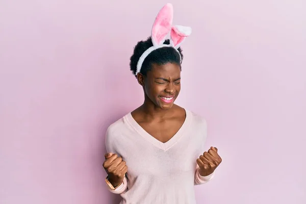 かわいいイースターバニーの耳を身に着けている若いアフリカ系アメリカ人の女の子は非常に満足し 腕を上げ 笑顔と成功のために叫んで勝者のジェスチャーを行うことに興奮しています お祝いのコンセプト — ストック写真