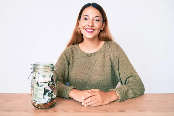 Masada duran genç esmer kadın kavanoz dolusu dolarla pozitif ve mutlu görünüyor. Kendine güvenen bir gülümsemeyle dişlerini gösteriyor. 