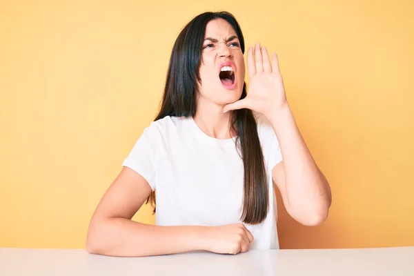 若い白人女性が机の上に座り 白いカジュアルなTシャツを着て大声で叫び 口の上で手を合わせて大声で叫ぶ コミュニケーションの概念 — ストック写真
