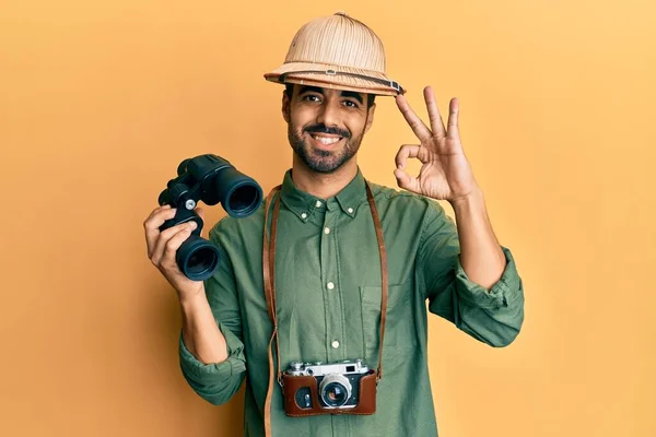 Young Hispanic Man Wearing Explorer Hat Looking Binoculars Doing Sign — Stock Photo, Image