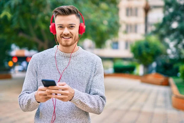 Parkta akıllı telefon ve kulaklık kullanarak gülümseyen beyaz bir genç..