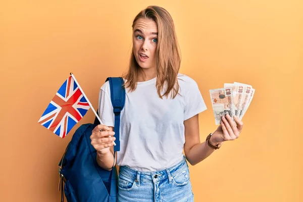 Beautiful blonde woman exchange student holding uk flag and pounds clueless and confused expression. doubt concept.