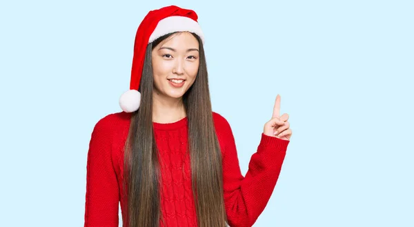 若い中国の女性が顔に大きな笑顔でクリスマスの帽子をかぶって カメラを見ている側に手指で指して — ストック写真