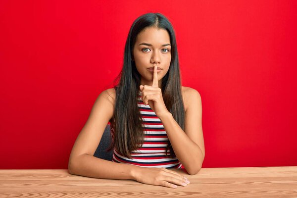 Красивая латиноамериканка в повседневной одежде сидит на столе и просит тишины с пальцем на губах. молчание и тайная концепция. 