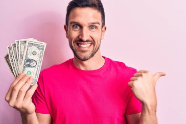 Soyutlanmış pembe arka planda bir deste banknot tutan yakışıklı bir adam başparmağıyla yan tarafa doğru gülümsüyor ve ağzı açık gülümsüyor.