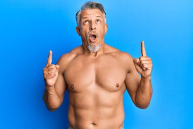 Orta yaşlı, gri saçlı, şaşkın ve şaşkın bir şekilde ayakta duran, parmaklarıyla işaret eden ve kollarını kaldıran adam.. 