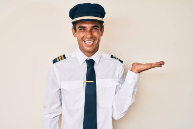 Uçak pilotu üniforması giyen, neşeli gülümseyen, avuç içiyle kameraya bakan İspanyol genç adam.. 