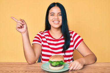 Lezzetli bir klasik burger yiyen Asyalı genç kız gülümsüyor. 
