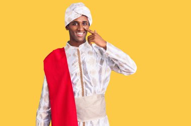 Gelenek giyinmiş Afrikalı yakışıklı bir adam Sherwani sari giysileri giyiyor. Parmakla yüzünü ve burnunu gösteriyor. Neşeli gülümsüyor. güzellik konsepti 