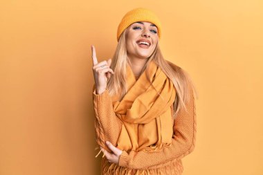 Yüzünde kocaman bir gülümseme olan yün kazak ve şapka giyen beyaz bir kadın kameraya el ve parmağıyla bakıyor.. 