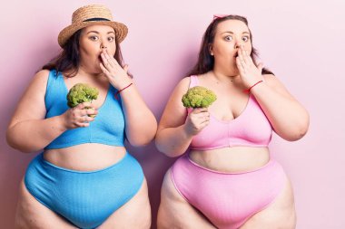 Bikini giyen, ağzı ağızla kaplayan brokoli tutan ve hata yapmaktan korkan genç ve büyük beden ikizler. şaşırmış ifade 