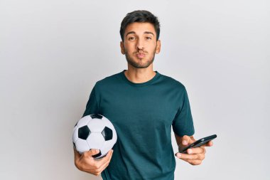 Genç yakışıklı adam futbol topu tutuyor ve akıllı telefon bahis uygulamasına bakıyor. Komik yüzüyle yanaklarını şişiriyor. Ağzı havayla şişer, havayı yakalar.. 