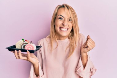 Güzel sarışın kadın elinde pasta şekerleriyle gururla bağırıyor zaferi kutluyor ve başarıyı kollarını kaldırarak kutluyor. 