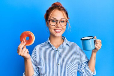 Kahvaltını çikolatalı çörek ve kahveyle yapan genç kızıl saçlı kadın yüzünde mutlu ve serin bir gülümsemeyle gülümsüyor. diş gösterme. 