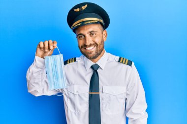 Sakallı, uçak pilotu üniforması giyen, güvenlik maskesi takan yakışıklı adam. Pozitif ve mutlu görünüyor. Kendine güvenen bir gülümsemeyle dişlerini gösteriyor. 