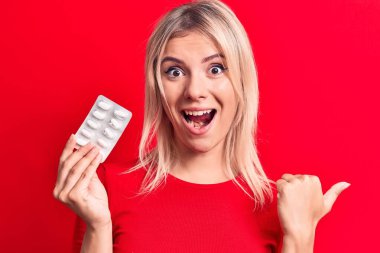 Sarışın, güzel bir kadın, elinde haplarla kırmızı arka planda dikiliyor. Başparmağıyla yan tarafa bakıyor, ağzı açık gülümsüyor.