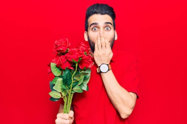 Sakallı, elinde gül buketi olan, ağzı elle kaplanmış, şok olmuş ve hata yapmaktan korkan genç, yakışıklı bir adam. şaşırmış ifade 