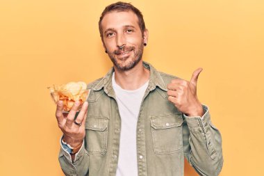 Elinde patates cipsi tutan yakışıklı adam mutlu ve pozitif gülümsüyor, başparmağıyla mükemmel ve onay işareti yapıyor. 
