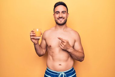 Tatilde sakallı, mayo giyen, bir bardak portakal suyu içen, mutlu, eli ve parmağı kenarda genç bir adam.