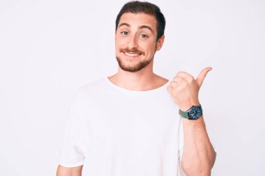 Gündelik beyaz tişört giyen, gülümseyen, gülümseyen ve başparmağıyla yan tarafı gösteren genç yakışıklı bir adam.. 