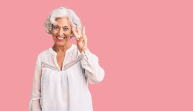 Kıdemli, gri saçlı, günlük kıyafetler giyen, üç numaralı parmaklarıyla işaret eden, kendinden emin ve mutlu bir şekilde gülümseyen kadın.. 