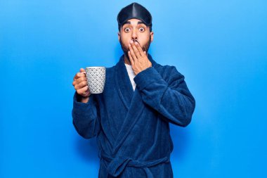 Uyku maskesi takan ve bornoz giyen, ağzı ağızla kaplanmış kahve içen, şok olmuş ve hata yapmaktan korkan genç bir İspanyol. şaşırmış ifade 