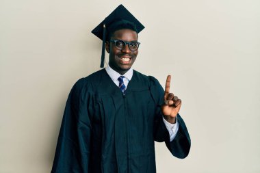 Mezuniyet şapkası ve tören cübbesi takan yakışıklı siyahi bir numaralı parmağıyla kendini beğenmiş ve mutlu gülümserken.. 