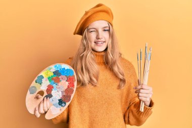 Güzel beyaz kız elinde boya fırçası ve ressam paleti ile kameraya seksi bir ifadeyle bakıyor, neşeli ve mutlu bir yüzle.. 