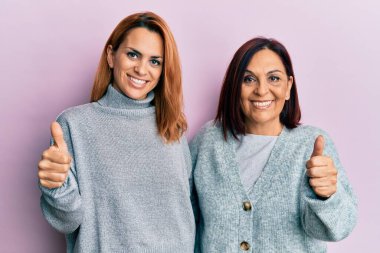Latin anne ve kızı gündelik giysiler giyerek mutlu bir şekilde el hareketi yapıyorlar. Kameraya bakıp başarıyı gösteren ifade. 