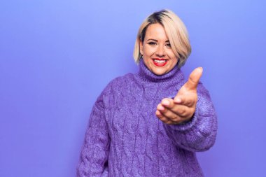 Güzel sarışın ve cüsseli bir kadın mor arka planda balıkçı yaka kazak giyiyor. Gülümsüyor, yardım eli uzatıyor ve kabul ediyor..