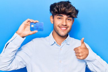 Genç İspanyol adamın elinde mutlu ve olumlu gülümseyen bir kredi kartı var. Başparmağıyla mükemmel ve onay işareti yapıyor. 