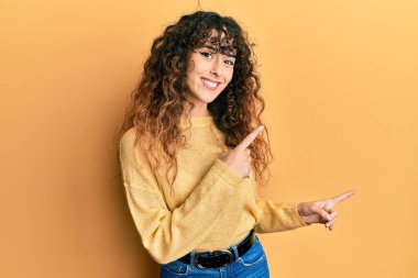 Gündelik kıyafetler giyen, gülümseyen ve kameraya bakan genç İspanyol kız iki eliyle ve parmaklarıyla kenarı işaret ediyor.. 