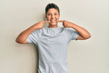 Gündelik gri tişört giyen, neşeli gülümseyen, parmaklarıyla ve ağzıyla işaret eden genç İspanyol çocuk. diş sağlığı kavramı. 