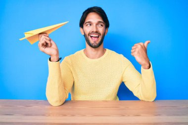 Yakışıklı İspanyol bir adam kağıt uçak tutuyor başparmağıyla yan tarafı işaret ediyor ağzı açık gülümsüyor. 
