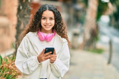 Güzel İspanyol kız şehirde akıllı telefon ve kulaklık kullanarak mutlu bir şekilde gülümsüyor..