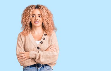 Kış kazağı giyen kıvırcık saçlı genç sarışın kadın kameraya bakarken kollarını kavuşturup gülümsüyor. pozitif kişi. 