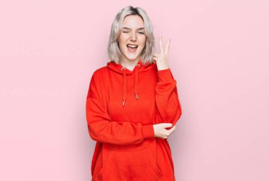 Sıradan kıyafetler giyen, gülümseyen, kameraya göz kırpan, zafer işareti yapan genç sarışın kız. İki numara.. 