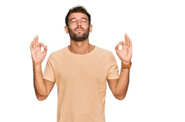 Sakallı, rahat bir tişört giyen, gözleri kapalı gülümseyen ve parmaklarıyla meditasyon yapan yakışıklı bir genç. yoga konsepti. 