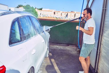 Orta yaşlı yakışıklı, günlük kıyafetler giyen ve mutlu gülümseyen güneş gözlüklü bir adam. Yüzünde gülümsemeyle ayakta durup su tabancası kullanarak araba yıkamak..