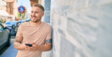 Genç İrlandalı mutlu bir şekilde gülümsüyor. Şehrin duvarına yaslanmış akıllı telefon kullanıyor..