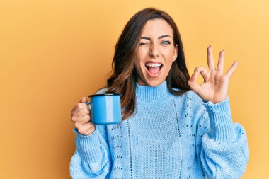 Genç esmer kadın elinde bir fincan kahve tutuyor parmaklarıyla imza atıyor, gülümseyerek mükemmel bir sembol oluşturuyor. 