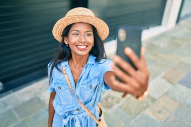 Genç, güzel Hintli bir kadın, yaz şapkası takıyor, gülümsüyor, mutlu bir şekilde şehrin akıllı telefonunda selfie çekiyor..