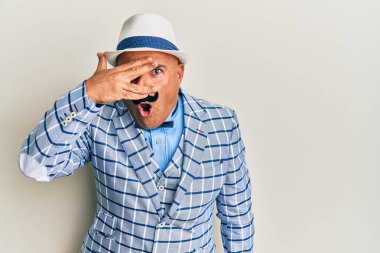 Olgun, orta doğulu bıyıklı, klasik ve şık moda tarzlı bir adam. Şok olmuş, yüzünü ve gözlerini eliyle kapatmış, parmaklarından korkuyla bakıyor. 