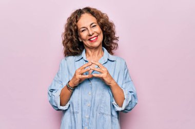 Orta yaşlı güzel bir kadın pembe arka plan üzerinde sıradan bir kot gömlek giyiyor ellerini birleştirip gülümsüyor rahat ve neşeli. Başarı ve iyimser