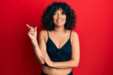 Kolsuz gömlekli, yüzünde kocaman bir gülümseme olan, eli ve parmağı kameraya bakan Latin kökenli genç bir kadın.. 