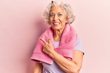 Kıdemli gri saçlı kadın spor giysisi ve havlu giyiyor. Gülümsüyor, eli ve parmağı yana doğru uzanıyor. 